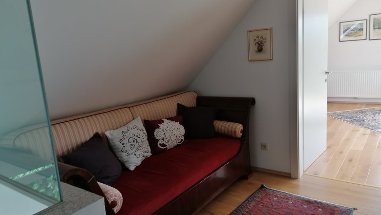 Sofa am Weg zum "Gartenzimmer", © Wiener Alpen Ein Sofa mit Kissen steht in einem Flur vor einer offenen Tür zu einem hellen Raum.