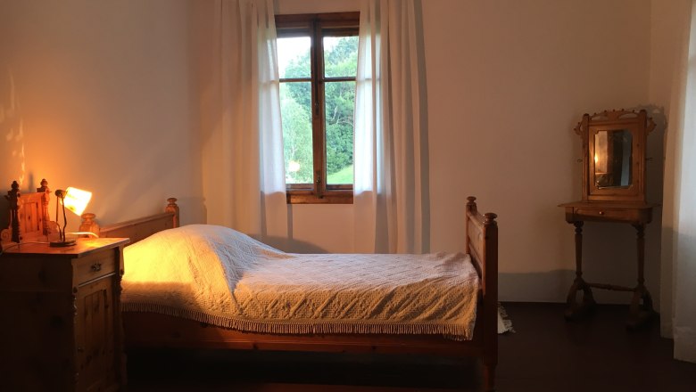 Einzelzimmer, © Riegelhof Ein rustikales Einzelzimmer mit Holzmöbeln, einem Bett, einem Nachttisch mit Lampe und einem Fenster mit weißen Vorhängen.