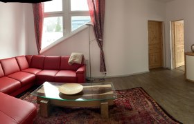 Wohnbereich, © Seidl Wohnzimmer mit rotem Ledersofa, Glastisch, Teppich und TV auf Holzschrank.