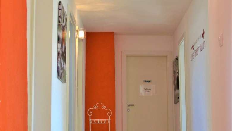 Aufgang, © Gluschitz-Goebel Ein Flur mit Holzboden, weißen Wänden und einer orangefarbenen Wand. An der Wand hängt ein Schild mit der Aufschrift 'Achtung Stufe'.