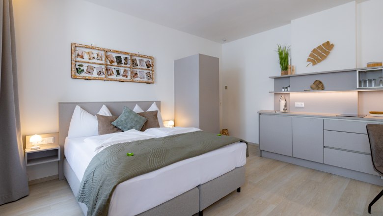 Apartment 2, © Atelier Fürtner-Ton Modernes Schlafzimmer mit Doppelbett, grauer Bettwäsche, Wanddekoration und kleiner Küchenzeile.