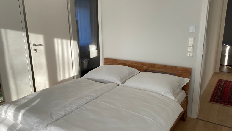 Schlafzimmer, © Wiener Alpen Ein Schlafzimmer mit einem Doppelbett, weißen Bettbezügen und Holzboden. Eine offene Tür führt zu einem Badezimmer.