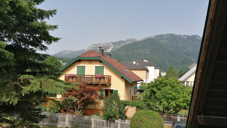 Blick vom großen Balkon (ca. 18m2), © Wiener Alpen Blick von einem Balkon auf ein gelbes Haus mit rotem Dach, umgeben von Bäumen und Bergen im Hintergrund.