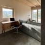 Arbeitsplatz, © Meinrad Hofer Ein moderner Arbeitsplatz mit einem Laptop auf einem Holztisch, einem schwarzen Stuhl und einem Sofa in einem Raum mit großen Fenstern und Waldblick.