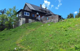Waldfreundehütte Obersberg, © Stefanie Gaulhofer Eine Berghütte auf einer grünen Wiese unter blauem Himmel.