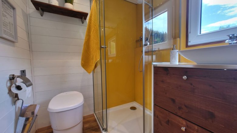 Badezimmer, © Villa to go Modernes Badezimmer mit gelber Duschwand, weißem WC, Holzregal und Fenster.