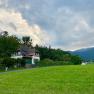 Orlet an der Rax, © Sofia Orlet Ein Haus im traditionellen Stil steht vor einem bewaldeten Hügel, umgeben von grünen Wiesen und Bergen im Hintergrund.
