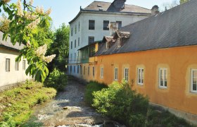 Brot- und Mühlenmuseum, © brot & mühle, T. Sommeregger, C. Dirnbacher Historische Gebäude an einem Bach mit blühenden Bäumen im Vordergrund.