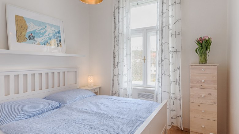 Top 3 - Schlafzimmer, © VP FeWo OG Helles Schlafzimmer mit weißem Bett, blauer Bettwäsche, Holzkommode und Fenster mit Vorhängen.