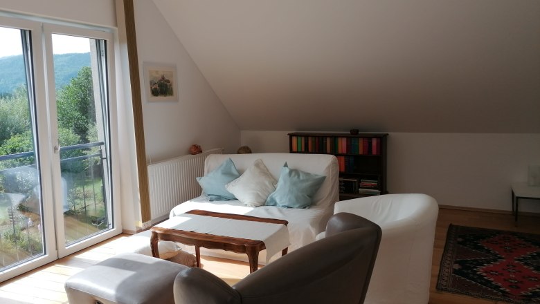 Wohnbereich im "Gartenzimmer", © Wiener Alpen Heller Wohnbereich mit Sofa, Sesseln, Bücherregal und großem Fenster mit Blick ins Grüne.
