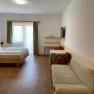 Familienzimmer, © Wiener Alpen Ein modernes Hotelzimmer mit Doppelbett, Sofa, Tisch und Stühlen. Ein Fernseher hängt an der Wand. Vorhänge an einem Fenster lassen Tageslicht herein.