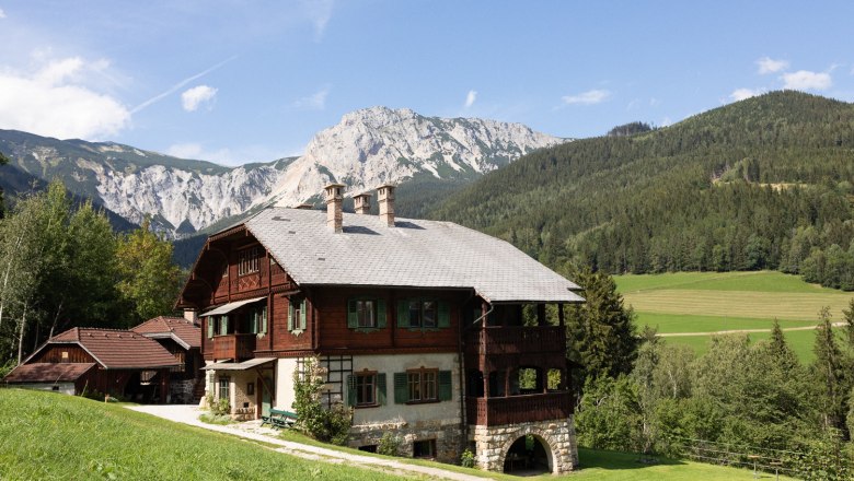 Der Riegelhof, © Wiener Alpen - Lierzer Ein traditionelles Holzhaus in einer bergigen Landschaft mit Wiesen und Wäldern im Hintergrund.