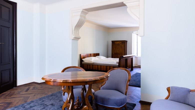 Zimmer, © Thalhof, Katrin Nusterer Ein elegantes Zimmer mit antiken Möbeln, darunter ein Holztisch, zwei Stühle und ein Bett im Hintergrund.