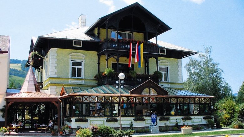 Café Reichenau, © Café Reichenau Ein traditionelles Gebäude mit Glasveranda und österreichischer Flagge, umgeben von Pflanzen.