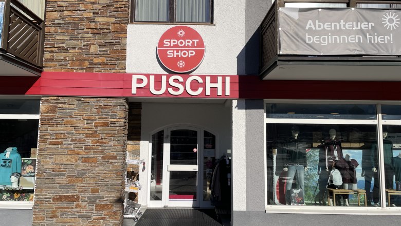 Sport-Puschi-Sportshop-Front, © Sport Puschi Eingangsbereich eines Sportgeschäfts mit der Aufschrift 'PUSCHI' und einem Schild 'SPORT SHOP'. Schaufenster mit Sportbekleidung und Ausrüstung.