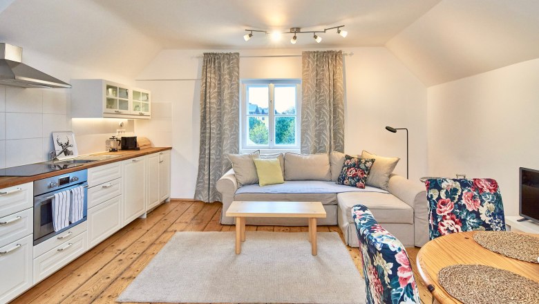 Top 7 - Wohnzimmer, © VP FeWo OG Gemütliches Wohnzimmer mit offener Küche, Holzboden, beiger Couch, floral gemusterten Stühlen und einem Fenster mit Vorhängen.