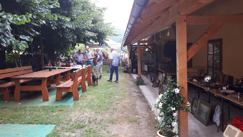 Gastgarten, © Thomas Lutz Ein Gastgarten mit Holztischen und Bänken, Menschen im Gespräch, ein Hund, und ein überdachter Bereich mit Flohmarkt.