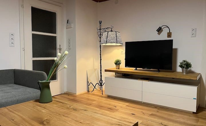 Wohnzimmer, © Anita Gallei Modernes Wohnzimmer mit Holzboden, grauem Sofa, TV auf weißem Schrank, Stehlampe und Dekorationen.