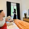 Joes Cozy Aparment, © Brunner Ein Mann sitzt auf einem Bett und schaut fern, während eine Frau am Fenster steht und Kaffee trinkt.