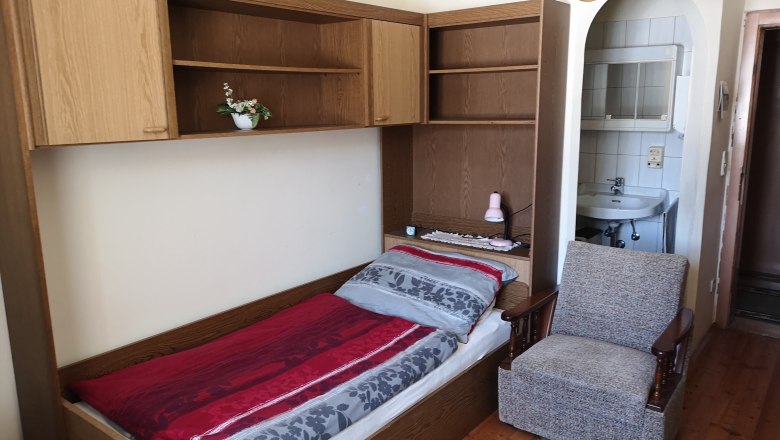 Einzelbett, © Gudrun Rath Einzelbett mit roter und grauer Bettwäsche, daneben ein Sessel und ein Waschbecken im Hintergrund.