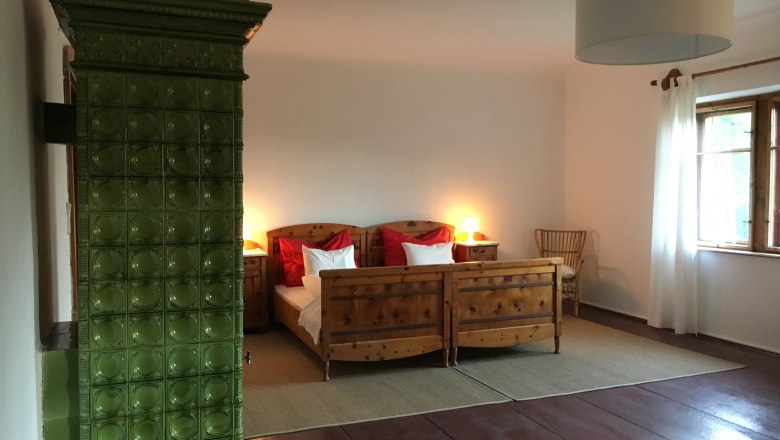 Doppelzimmer, © Riegelhof Ein gemütliches Doppelzimmer mit Holzmöbeln, einem grünen Kachelofen und einem großen Fenster mit weißen Vorhängen.