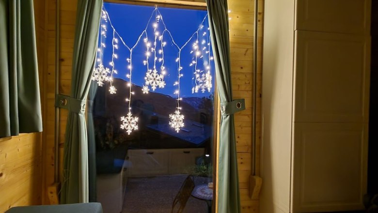 Wir vermieten auch im Winter!, © Villa to go Fenster mit Vorhängen und Schneeflocken-Lichterkette, zwei Kerzen auf der Fensterbank.