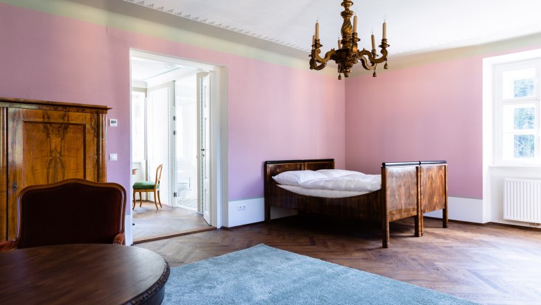 Zimmer, © Thalhof, Katrin Nusterer Ein elegantes Zimmer mit rosa Wänden, einem Holzbett, einem Kronleuchter und einem Teppich auf Holzboden.