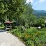 Garten, © Konstantin Hübner Ein gepflegter Garten mit Kiesweg, Blumenbeeten, einem Pavillon und einer Liege. Im Hintergrund sind Bäume und Berge zu sehen.