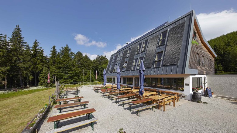 Naturfreundehaus Knofeleben, © Wiener Alpen / Bene Croy Ein modernes Gebäude mit Solarpanelen und Biergarten im Freien, umgeben von Wald.