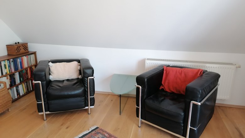 Wohnbereich im "Balkonzimmer", © Wiener Alpen Zwei schwarze Ledersessel mit Kissen, ein Glas-Beistelltisch, Bücherregal und Teppich in einem Wohnbereich mit Dachschräge.