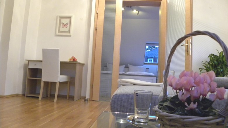 Zimmerbeispiel, © Thomas Baldauf Innenraum mit Schreibtisch, Stuhl, Blumen im Korb und Blick ins Schlafzimmer.