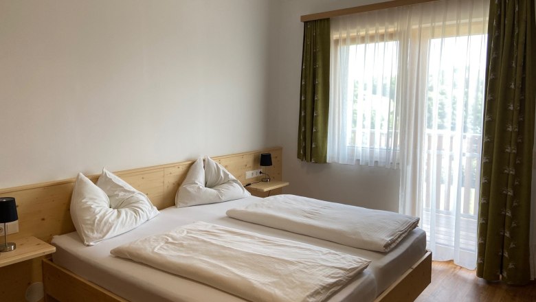 Familienzimmer, © Wiener Alpen Ein helles Schlafzimmer mit einem Doppelbett, weißen Bettdecken und Kissen. Ein Fenster mit grünen Vorhängen lässt Tageslicht herein.