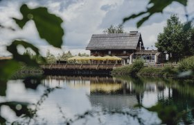 Am Fuße des Hirschenkogels (Zauberberg Semmering), © Niederösterreich Werbung/Rita Newman Ein rustikales Holzhaus mit gelben Sonnenschirmen spiegelt sich in einem ruhigen See, umgeben von grüner Natur.