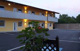 Aussenansicht, © Alexandra Heiling-Niegl Außenansicht eines zweistöckigen Gebäudes mit gelber Fassade und beleuchtetem Balkon, davor ein geparkter roter Wagen und Pflanzen im Vordergrund.