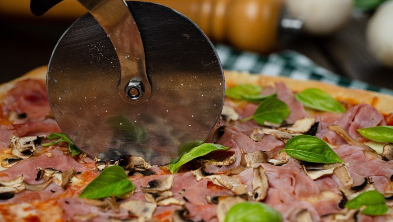 Pizzaspezialitäten, © cervodoro Nahaufnahme einer Pizza mit Schinken, Pilzen und Basilikum, die mit einem Pizzaschneider geschnitten wird.