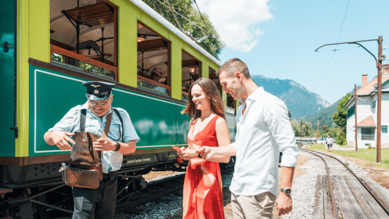 Höllentalbahn, © Österreich Werbung/Stefan Strasser Ein Schaffner in Uniform zeigt einem Paar vor einem grünen Zug etwas aus seiner Tasche. Im Hintergrund sind Gleise und Berge zu sehen.