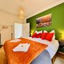 Joes Coxy Apartment, © Brunner Gemütliches Schlafzimmer mit grün gestrichener Wand, Doppelbett mit roten Kissen, oranger Decke und Landschaftsbild. Hängelampe und Kleiderständer vorhanden.