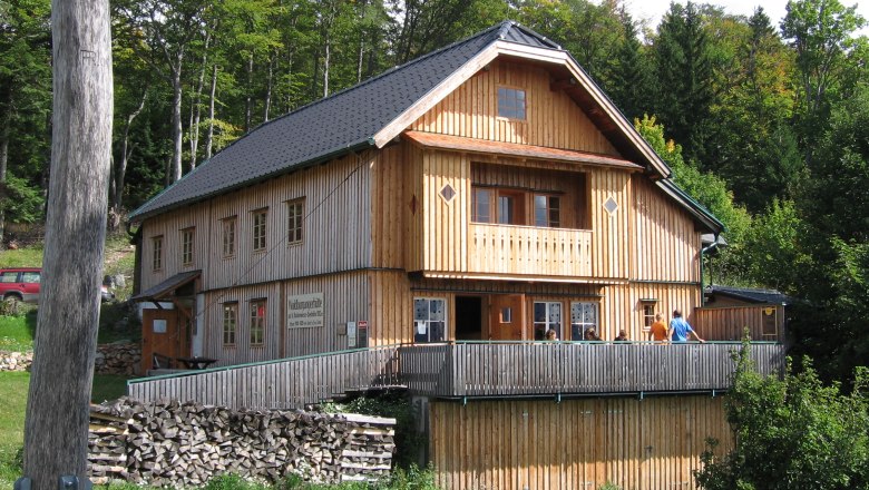 Waldburgangerhütte, © Maria Stroebl Eine Holzhütte im Wald mit Terrasse und Personen darauf.