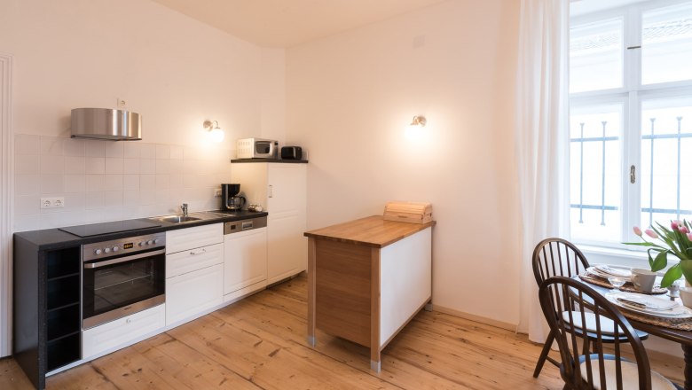 Top 2 - Küche, © VP FeWo OG Moderne Küche mit Holzboden, weißer Einrichtung und kleinem Esstisch am Fenster.