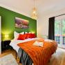 Joes Coxy Apartment, © Brunner Gemütliches Schlafzimmer mit grün gestrichener Wand, Doppelbett mit orangefarbenen Decken und Kissen, Lampe und Balkon.