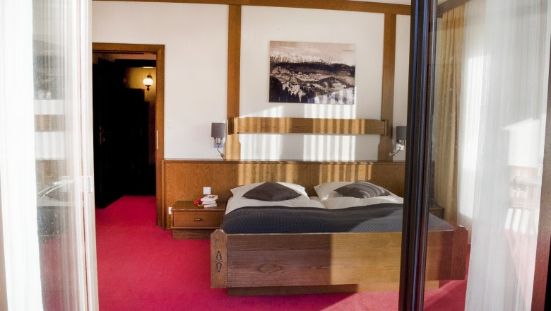 Zimmer, © Doris Seebacher Ein gemütliches Schlafzimmer mit einem Doppelbett, Holzmöbeln und einem Bild an der Wand. Der Boden ist mit einem roten Teppich ausgelegt.