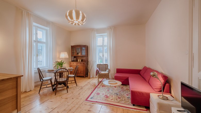 Top 2 - Wohnzimmer, © VP FeWo OG Gemütliches Wohnzimmer mit rotem Sofa, Holzboden und Esstisch.