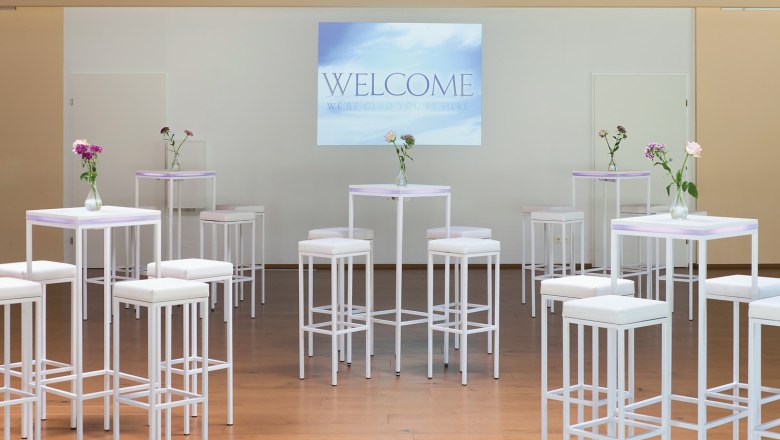 Seminar im Parkhotel Hirschwang, © missionINGE.com Seminarraum mit weißen Stehtischen und Hockern, Blumenvasen und einer Leinwand mit der Aufschrift 'Welcome'.
