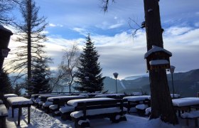 Terrasse Speckbacher Hütte, © Karin Stranz Verschneite Terrasse mit Holzbänken und Tischen, umgeben von Bäumen und Bergen im Hintergrund.