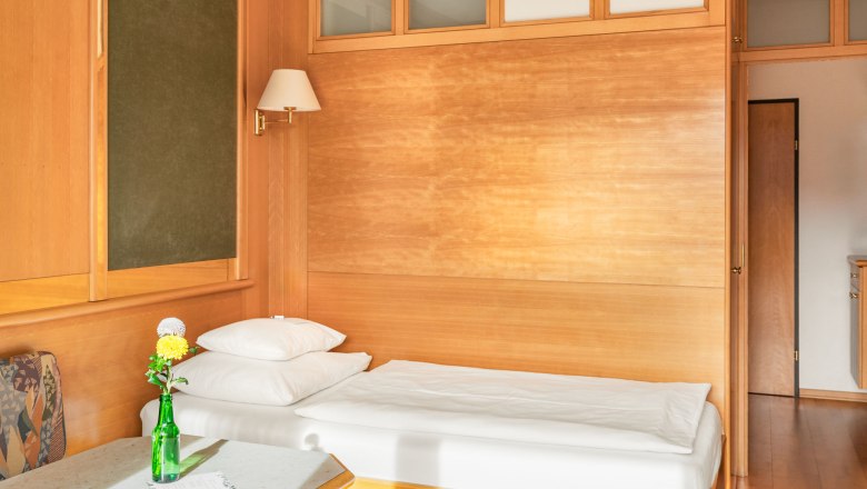 Einzelzimmer, © Kristina Polleres Ein einfach eingerichtetes Zimmer mit einem Einzelbett, Holzwänden und einem Tisch mit einer Vase und Blumen.