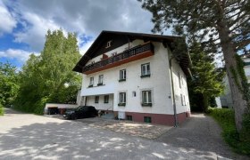 Hausansicht, © Brunner Zweistöckiges Haus mit weißer Fassade und dunklem Dach, umgeben von Bäumen und einem gepflasterten Hof.