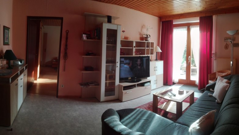 Wohnzimmer, © Kesper Gemütliches Wohnzimmer mit Sofa, Fernseher, Regal und Glastür mit roten Vorhängen.
