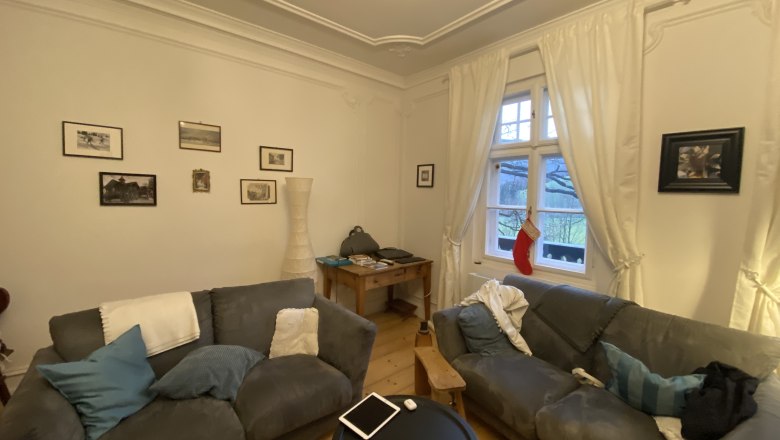 753e5cc4-a3ed-4959-8b93-e84a4e92cea7, © Martin Sturm Gemütliches Wohnzimmer mit zwei grauen Sofas, einem runden Tisch, Bildern an der Wand und einem Fenster mit Vorhängen und Weihnachtsstrumpf.