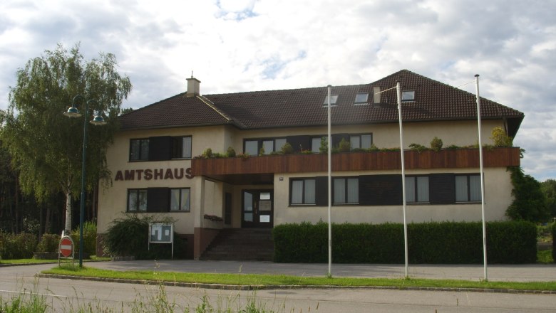 Amtshaus Natschbach-Loipersbach, © Gemeinde Natschbach-Loipersbach Das Amtshaus Natschbach-Loipersbach mit braunem Dach und Balkon, umgeben von Bäumen und Fahnenmasten.