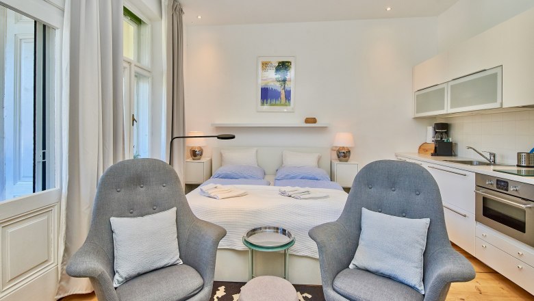 Top 4, © VP FeWo OG Modernes Studio-Apartment mit Doppelbett, zwei grauen Sesseln, Küchenzeile und großen Fenstern.